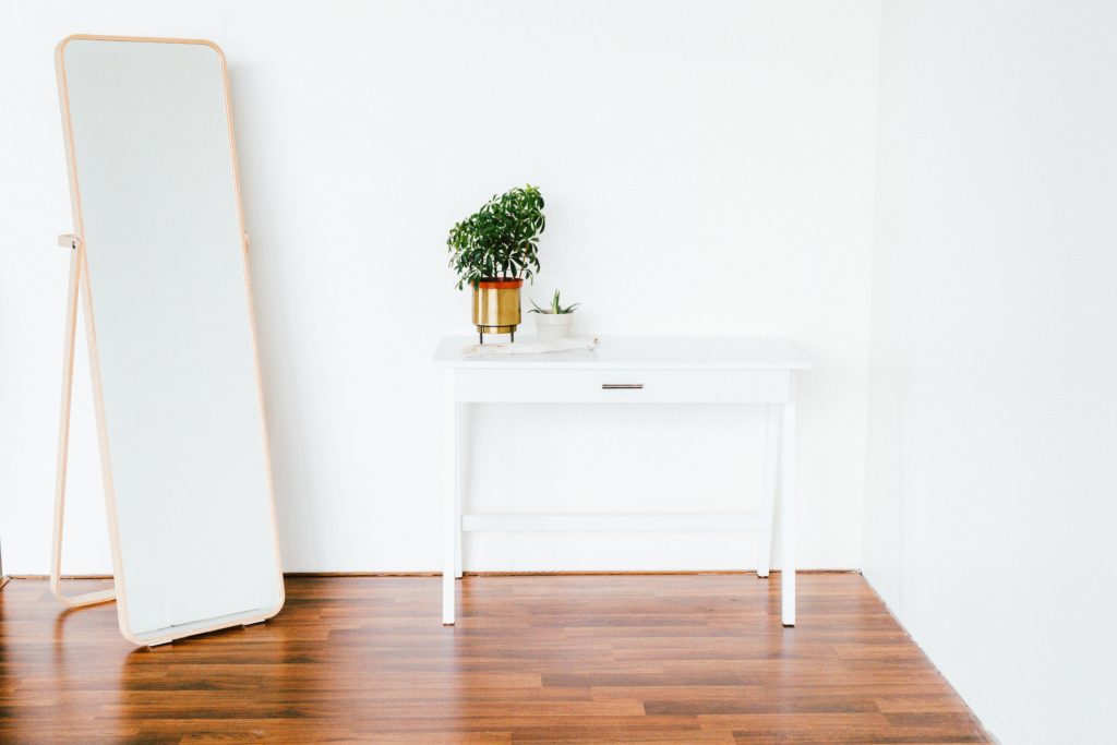 Une entrée minimaliste : une console blanche et un miroir sur pieds. 
Une plante dans un cache pot doré posé sur le meuble.